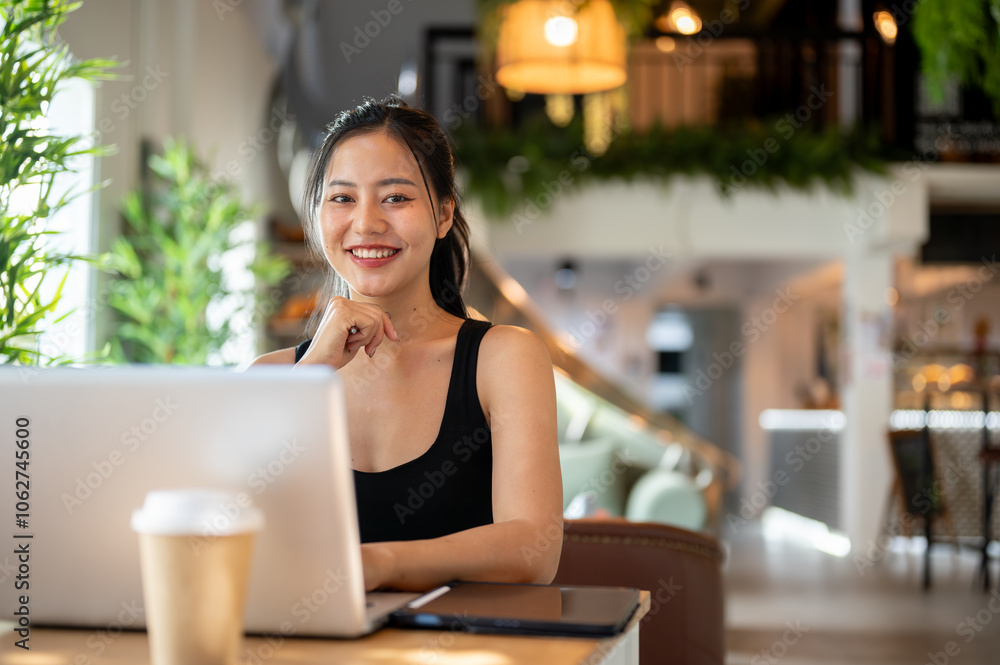 An Asian woman sitting at an indoor table in a coffee shop with her laptop, smiling at the camera. An Asian woman sitting at an indoor table in a coffee shop with her laptop, smiling at the camera.