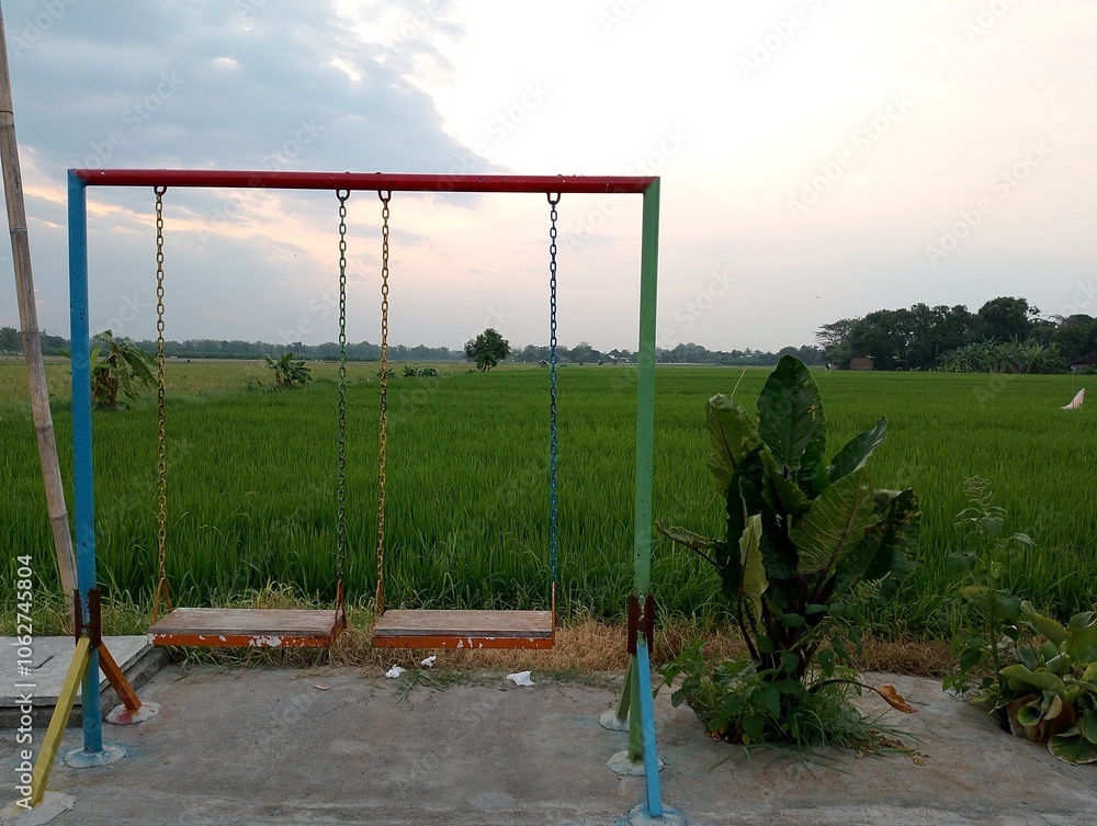 Fototapeta premium A single swing stands in the middle of a lush green rice field, creating a serene and picturesque scene.