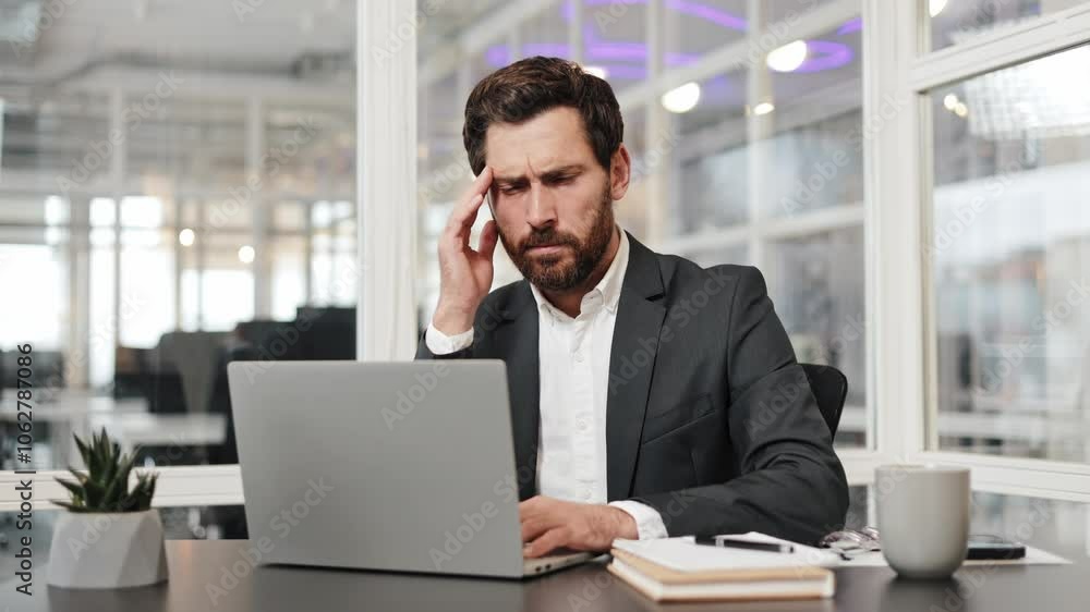 Tired businessman in a suit sits at his desk in the office, experiencing a headache while working on his laptop. Manager rubs his temples as he reads the screen, expressing fatigue and stress. Tired businessman in a suit sits at his desk in the office, experiencing a headache while working on his laptop. Manager rubs his temples as he reads the screen, expressing fatigue and stress.