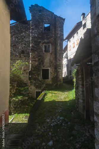 Un vicolo del borgo di Erto presso la diga del Vajont con la sua pavimentazione ad acciottolato coperta di erba passa fra alcune case diroccate in una giornata di sole photo