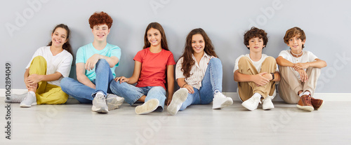 Portrait of a smiling school children looking cheerful at the camera on gray wall background. Confident boys and girls in casual clothes sitting in a row on the floor. Kids and education concept.