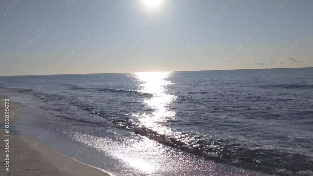The image captures a tranquil ocean scene with gentle waves washing up on a sandy shore. Sunlight reflects off the water, creating a bright, shimmering path on the sea’s surface, under a clear sky. The image captures a tranquil ocean scene with gentle waves washing up on a sandy shore. Sunlight reflects off the water, creating a bright, shimmering path on the sea’s surface, under a clear sky.