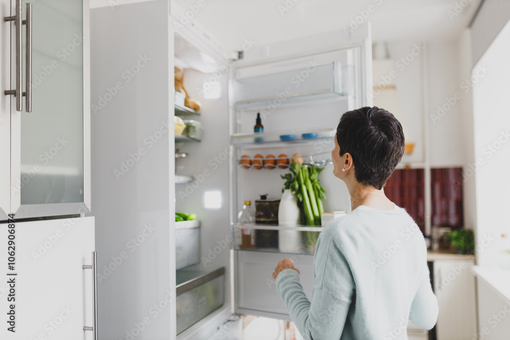 Back view of brunette woman with short hair standing in front of opened refrigerator full of food products, choosing ingredients to cook nutritious healthy breakfast before going to work Back view of brunette woman with short hair standing in front of opened refrigerator full of food products, choosing ingredients to cook nutritious healthy breakfast before going to work