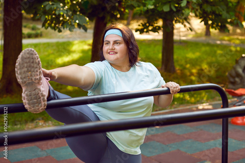 Happy beautiful smiling fat plump stout overweight adult woman in headband doing leg stretching exercise by high meal bar during fitness body workout on urban sports ground among green park trees photo