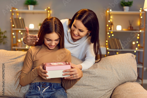 Happy cheerful young mother congratulating her smiling teenage child girl on holiday sitting on sofa at home. Joyful woman making daughter surprise giving her present gift box. Family concept. photo