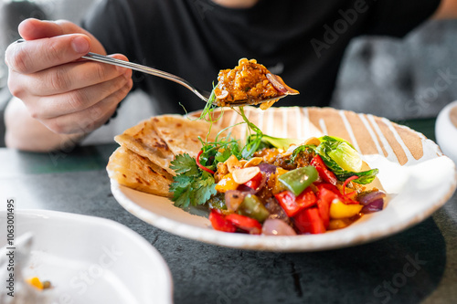 A person enjoying a colorful meal with fresh vegetables and flatbread on a wooden table. The vibrant dish includes tomatoes, cucumbers, and greens, perfect for culinary and healthy eating themes. photo