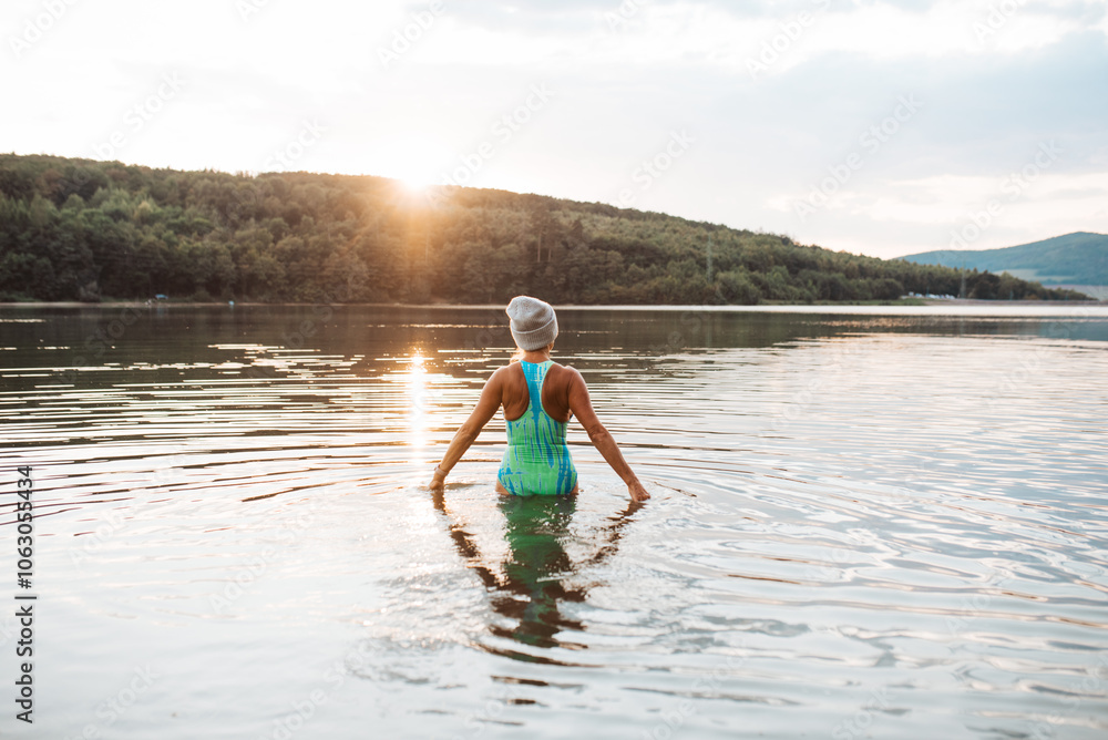 Cold water swimming for elderly women. Senior sporty women standing in lake during cold evening, looking at setting sun.