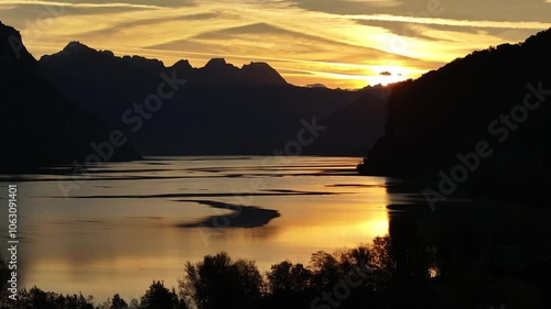 Camera slowly pushes toward Walensee lake with golden sun rays reflecting off water, ajestic Swiss alps and warm yellow sky at sunrise in Quinten, Mols, Walenstadt, Switzerland. photo