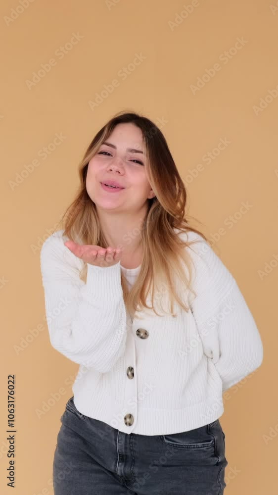 Positive young adult Caucasian woman sending an air kiss, standing on beige background. Gesture of tenderness and affection. Vertical video.
