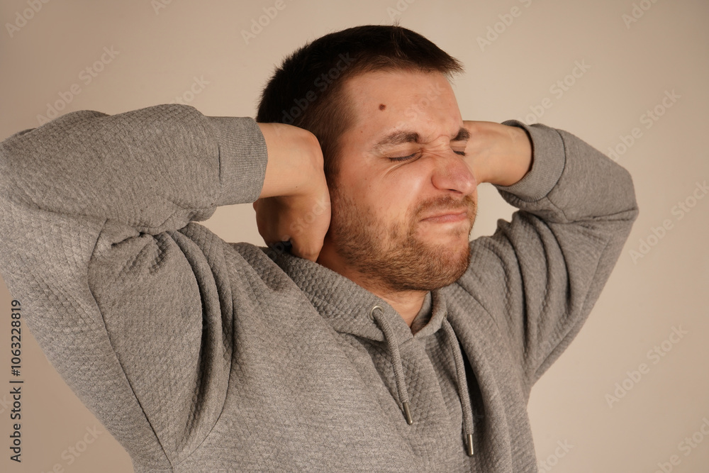 Fototapeta premium Portrait of young man closing his ears by hands and keeping his eyes closed while isolated on grey. Photo of desperate annoyed bearded man keeps hands on head and closes eyes, feels depressed.