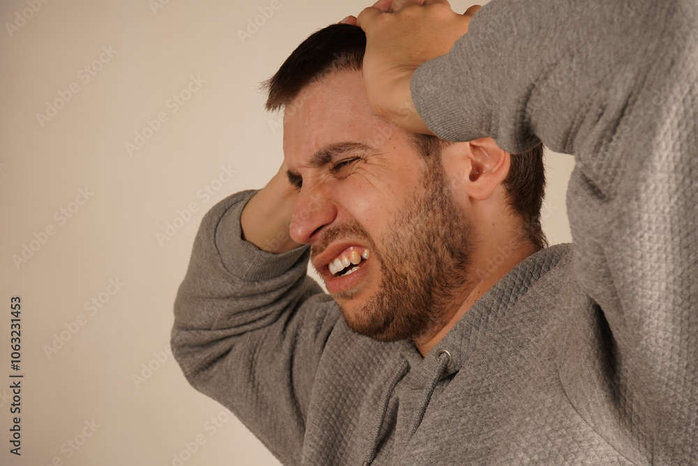 Fototapeta premium Man in depression. Portrait of young guy closing his ears by hands and keeping his eyes closed while isolated on grey. Photo of desperate annoyed bearded man keeps hands on head. Man with headache.