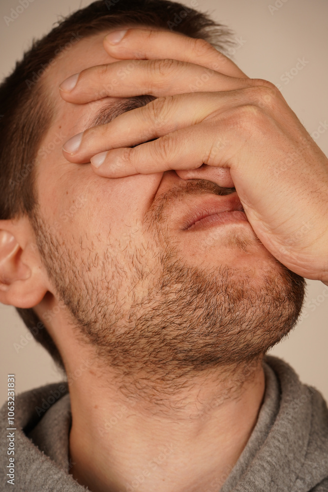 Obraz premium Facepalm sign. Portrait of desperate man with beard in hoodie covering face with hand, feeling sorry and blaming himself for the mistake. indoor studio shot isolated on grey background.