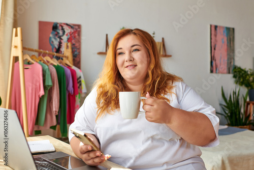 A plus size woman smiles softly while sipping coffee and using her phone in a stylish, serene space.