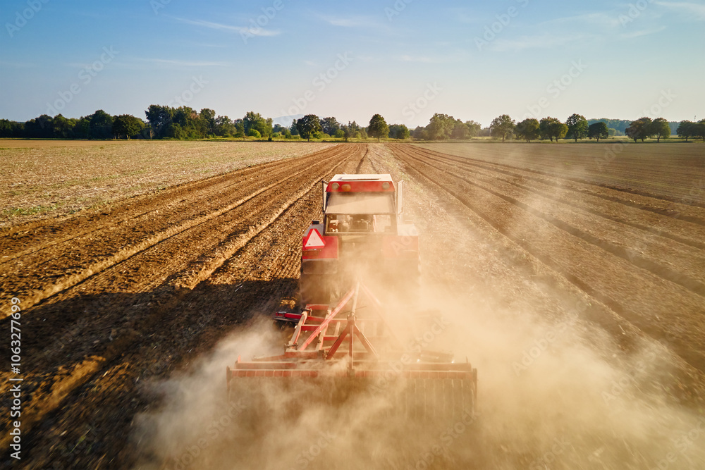 Fototapeta premium Red tractor working in agricultural field, cultivating and plowing dry soil, aerial view. Agribusiness concept. Small farming concept