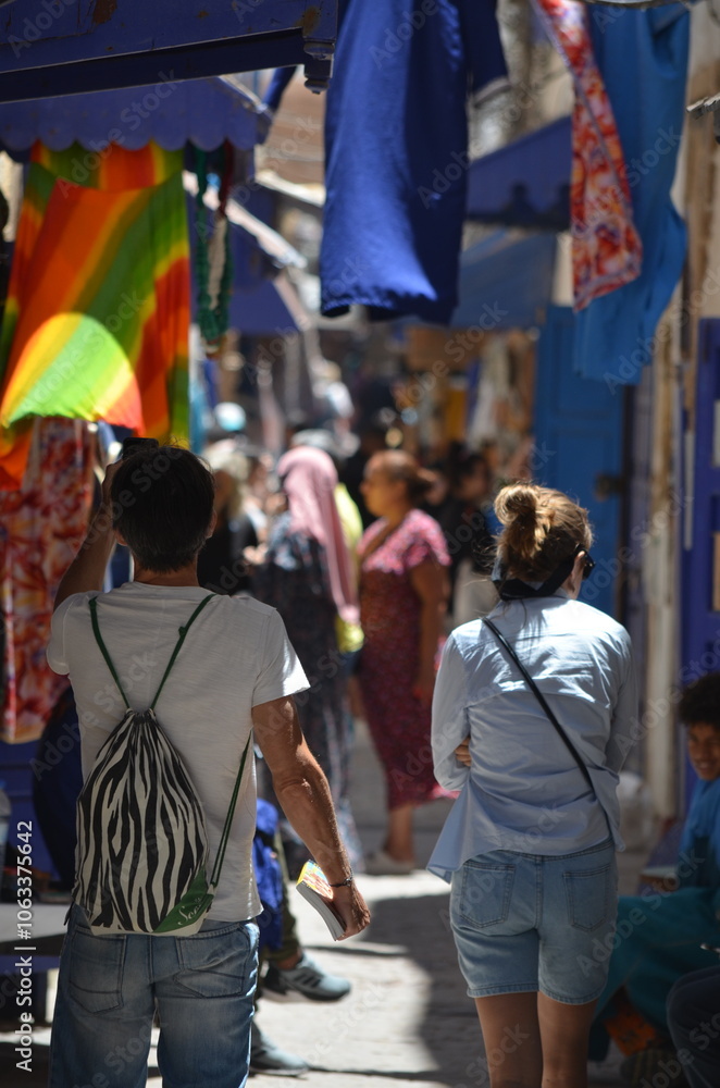 Obraz premium Tourists walking through a narrow, vibrant alleyway in Essaouira's Medina, with colorful textiles hanging from the blue awnings and locals sitting along the street 