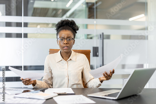 Young African American businesswoman holding papers looking confused at desk. Office environment with laptop, documents suggest stress, decision making in business setting.