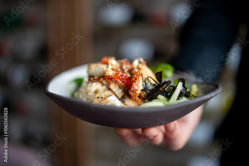 Fototapeta Naklejka Na Ścianę i Meble -  A close-up of a hand holding a bowl of Asian cuisine, featuring rice and vegetables, garnished with sesame seeds and herbs. vibrant colors and fresh ingredients create an appetizing and healthy meal.