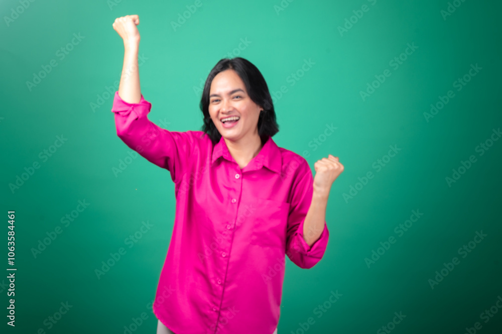 An Asian woman in a pink shirt celebrates with a joyful expression, raising her fists in excitement. She stands against a green background, exuding a sense of achievement and happiness An Asian woman in a pink shirt celebrates with a joyful expression, raising her fists in excitement. She stands against a green background, exuding a sense of achievement and happiness