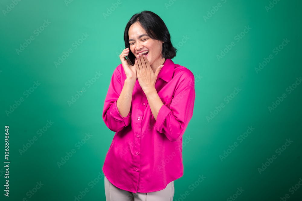 An Asian woman wearing a bright pink shirt poses while talking on her smartphone. She has one hand on her hip and a cheerful expression on her face. An Asian woman wearing a bright pink shirt poses while talking on her smartphone. She has one hand on her hip and a cheerful expression on her face.