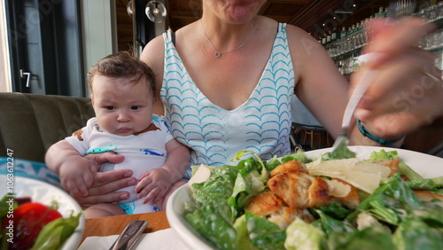 Mother holding baby while eating a fresh salad at a restaurant, baby looking curiously at the food, cozy family mealtime scene with mother multitasking and caring for the baby