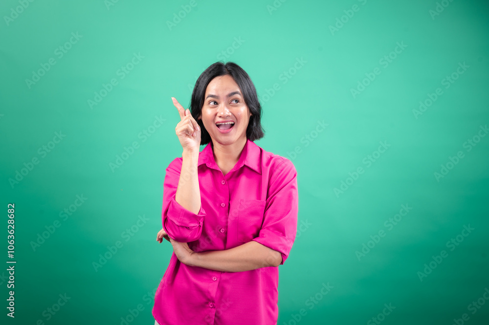 An Asian woman in a vibrant pink shirt stands against a green background, looking cheerful and pointing upwards with one hand, as if indicating a bright idea or something exciting An Asian woman in a vibrant pink shirt stands against a green background, looking cheerful and pointing upwards with one hand, as if indicating a bright idea or something exciting