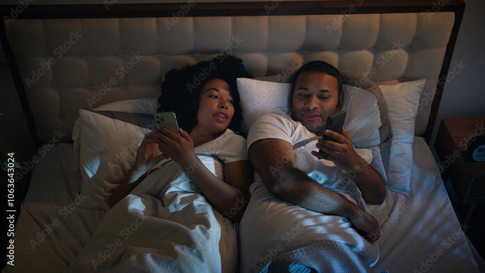 Obraz premium African American couple in bed at night late time in game bedroom relaxing using two phones chatting in mobile app scrolling social media smartphones woman man wife husband talking discuss news family