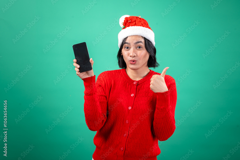 An Asian woman in a red sweater and Santa hat winks and gives a thumbs-up while holding a smartphone against a green background, expressing excitement and holiday cheer. An Asian woman in a red sweater and Santa hat winks and gives a thumbs-up while holding a smartphone against a green background, expressing excitement and holiday cheer.
