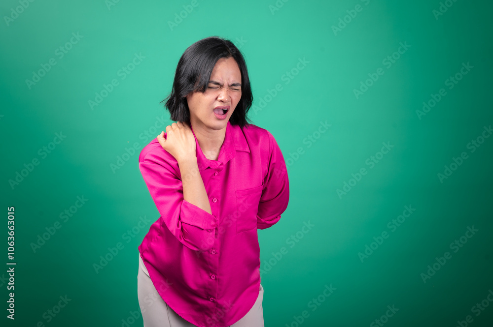 Fototapeta premium An Asian woman wearing a bright pink shirt against a green background is holding her shoulder with one hand, expressing pain or discomfort, with her face showing a pained expression