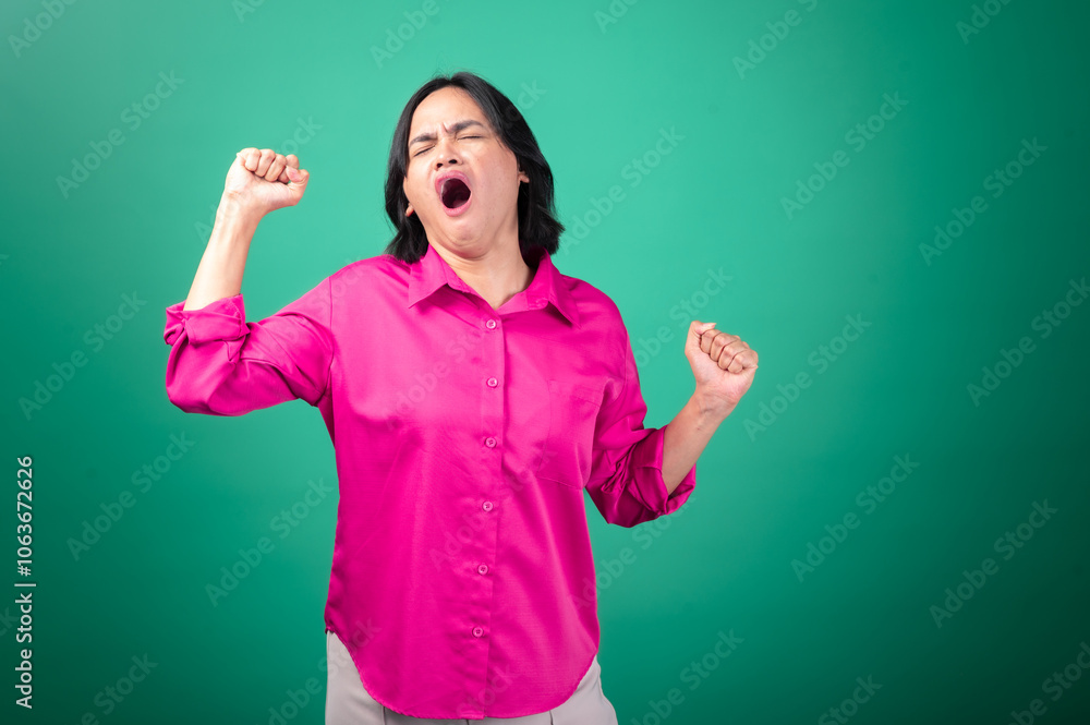 Asian woman in a bright pink shirt against a green background appears to be yawning, with arms stretched out in a gesture of tiredness or fatigue. Her facial expression body language sleepiness. Asian woman in a bright pink shirt against a green background appears to be yawning, with arms stretched out in a gesture of tiredness or fatigue. Her facial expression body language sleepiness.