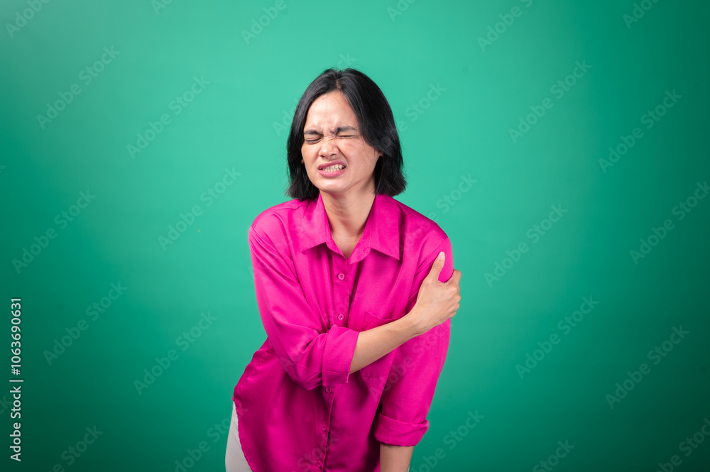 Obraz premium An Asian woman wearing a bright pink shirt against a green background is holding her shoulder with one hand, expressing pain or discomfort, with her face showing a pained expression