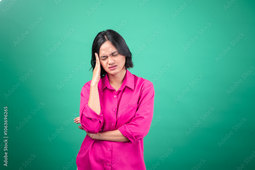 An Asian woman in a bright pink shirt against a green background holds her hand to her head, eyes closed, expressing a feeling of headache or stress. An Asian woman in a bright pink shirt against a green background holds her hand to her head, eyes closed, expressing a feeling of headache or stress.