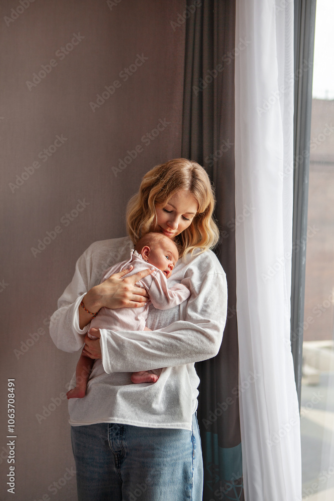 Fototapeta premium Young beautiful mother holds newborn baby while standing at home near window. tender moment with baby dressed in soft pink And mom in light clothes. Natural light, peaceful and intimate atmosphere