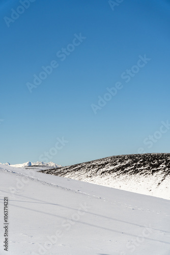 Obraz na płótnie A vertical perspective of a snowy hill featuring rocky textures against a bright blue sky, highlighting the beauty of the winter landscape. w salonie