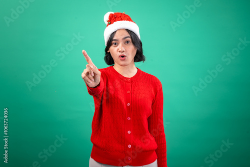 An Asian woman in a red sweater and Santa hat stands against a green background, pointing her finger forward with a serious expression, creating a direct and attention-grabbing holiday-themed