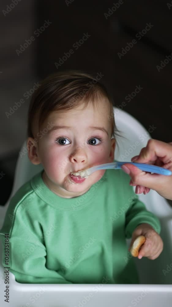 Nourishing the little toddler sitting in the feeding table. High angle view. Baby holds a bagel in left hand and looks up at camera. Dark blurred backdrop. Vertical video Nourishing the little toddler sitting in the feeding table. High angle view. Baby holds a bagel in left hand and looks up at camera. Dark blurred backdrop. Vertical video