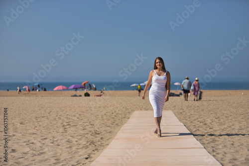 Fotografi Young beautiful woman in a white dress and heels parading on a wooden walkway on the beach
