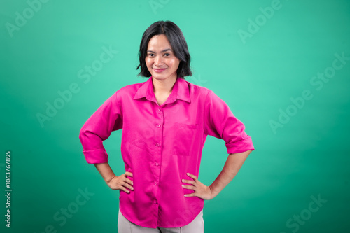 An Asian woman in a bright pink button-up shirt stands confidently with her hands on her hips against a solid green background, projecting a friendly and assertive expression photo