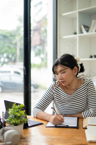 Intelligent young woman having online lessons on laptop, drinking coffee and taking notes,