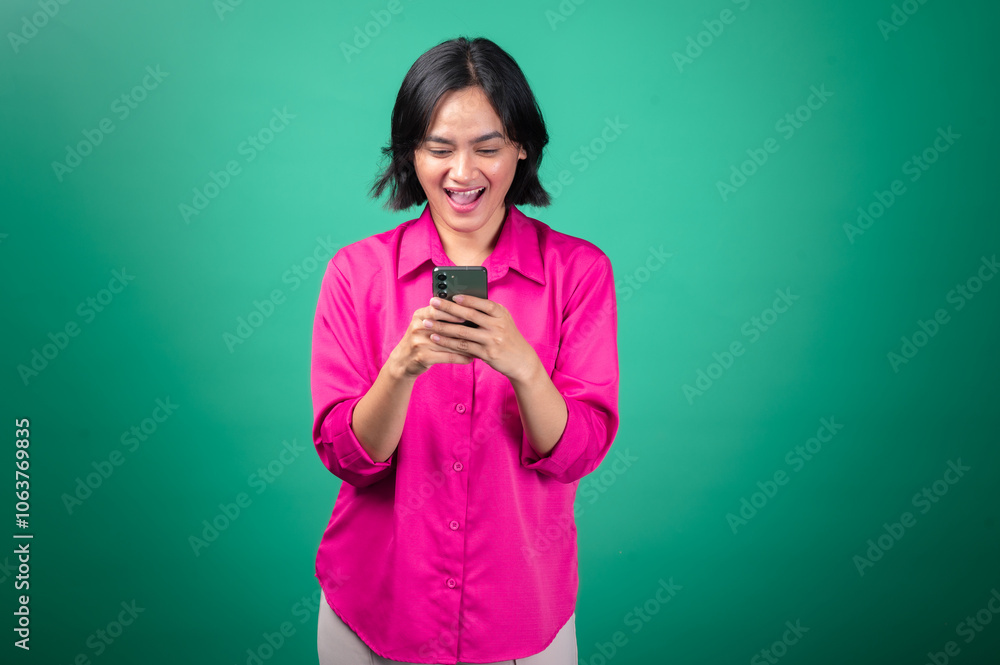 An Asian woman in a bright pink shirt smiles while looking down at her smartphone, holding it with both hands as if interacting with a game or reading something exciting. Her expression is cheerful An Asian woman in a bright pink shirt smiles while looking down at her smartphone, holding it with both hands as if interacting with a game or reading something exciting. Her expression is cheerful