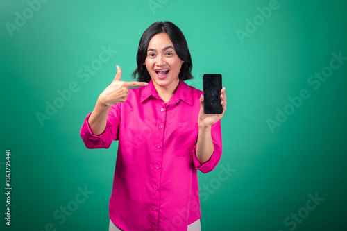 An Asian woman in a bright pink shirt holds up a smartphone with a big smile, pointing excitedly at the screen against a green background.
