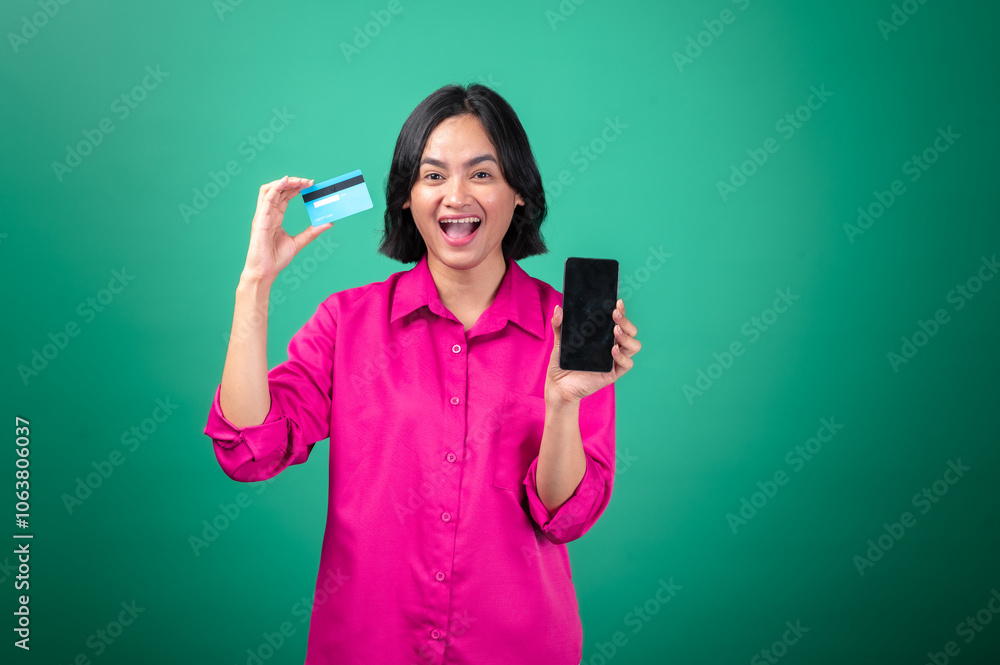 An Asian woman in a pink shirt against a green background, holding a credit card in one hand and a smartphone in the other. She smiles enthusiastically, symbolizing online shopping or digital payment An Asian woman in a pink shirt against a green background, holding a credit card in one hand and a smartphone in the other. She smiles enthusiastically, symbolizing online shopping or digital payment