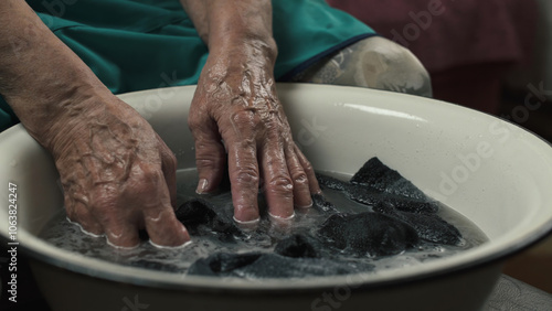 Elderly woman washing socks in white enamel basin while sitting on sofa.Close-up