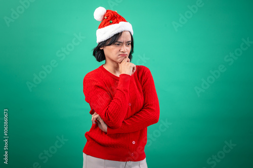 An Asian woman in a red sweater and Santa hat poses with a thoughtful expression, resting her chin on her hand, against a green background, creating a humorous and festive holiday mood