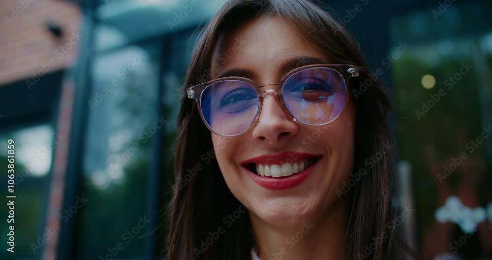 Very close facial view of Caucasian woman with round glasses looking at something on side. Glancing directly at camera. Showing warm smile on face. Trees visible in tall windows in background. Very close facial view of Caucasian woman with round glasses looking at something on side. Glancing directly at camera. Showing warm smile on face. Trees visible in tall windows in background.