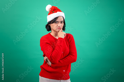 An Asian woman in a red sweater and Santa hat poses with a thoughtful expression, resting her chin on her hand, against a green background, creating a humorous and festive holiday mood photo