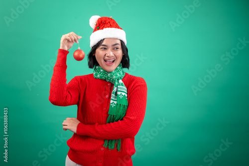 An Asian woman wearing a Santa hat, a red sweater, and a green holiday scarf smiles while holding a red Christmas ornament against a green background, radiating festive cheer