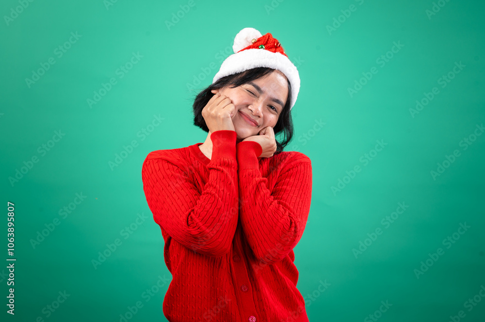 An Asian woman in a red sweater and Santa hat, standing against a green background, holds her hands near her cheeks, smiling sweetly with a playful and charming expression. She exudes holiday An Asian woman in a red sweater and Santa hat, standing against a green background, holds her hands near her cheeks, smiling sweetly with a playful and charming expression. She exudes holiday