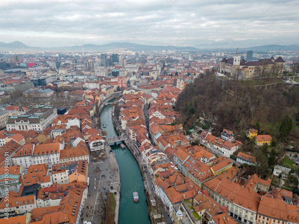 Naklejka premium Panorama of Ljubljana city. Aerial view above Ljubljanica river with river cruise boats towards city center. Historical town with old architecture and the castle on the hill. Cloudy day in the winter.