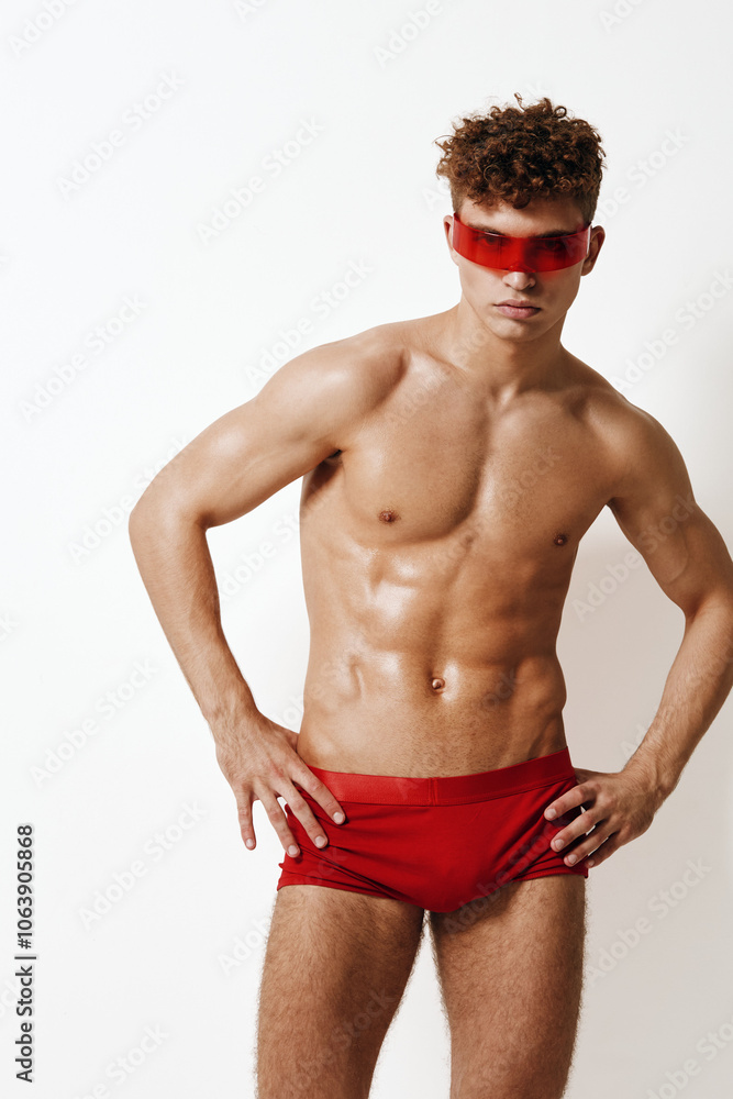 Obraz premium Confident young man in red swimwear and stylish glasses posing against a white background, showcasing fitness and modern fashion.