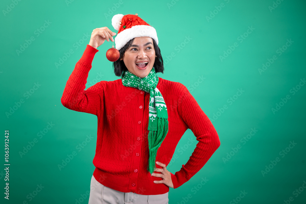 An Asian woman wearing a Santa hat, a red sweater, and a green holiday scarf smiles while holding a red Christmas ornament against a green background, radiating festive cheer An Asian woman wearing a Santa hat, a red sweater, and a green holiday scarf smiles while holding a red Christmas ornament against a green background, radiating festive cheer